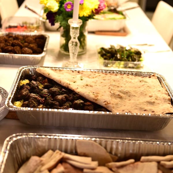 Mediterranean catering spread with meat, rice, and flatbread in foil pans on a white tablecloth.