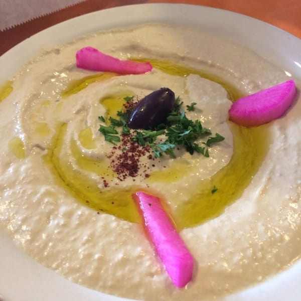Creamy hummus drizzled with olive oil, garnished with parsley, sumac, an olive, and pink pickled turnips.